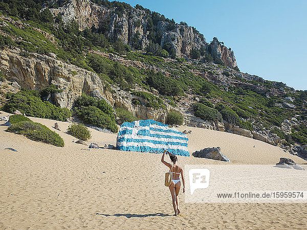 Rückansicht einer jungen Frau im Bikini  die am Sandstrand entlang auf einen mit der griechischen Flagge bemalten Felsbrocken zuläuft.