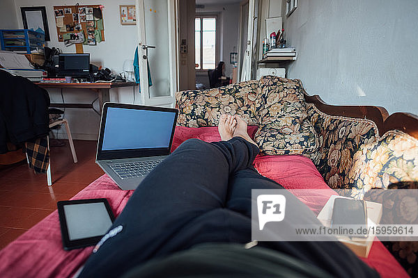 Mann auf Wohnzimmersofa liegend  mit digitalem Tablet  Laptop und Mobiltelefon  während er sich während der Corona-Krise selbst isoliert.