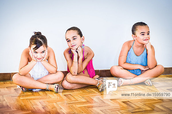 Drei junge Mädchen im Tanzkurs  geschminkt und in Sommerkleidern auf Parkett sitzend.