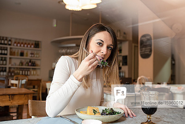 Eine Frau sitzt an einem Café-Tisch und isst eine Schale mit veganem Essen  Gemüse auf der Gabel.