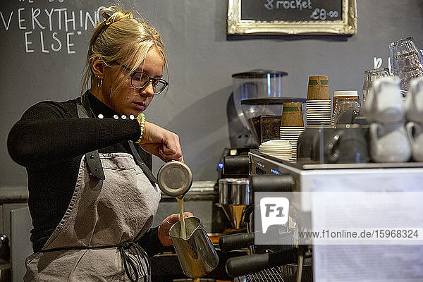 Blonde Frau mit Brille und Schürze  die an einer Espressomaschine in einem Café steht und Milch in eine Metallkanne gießt.