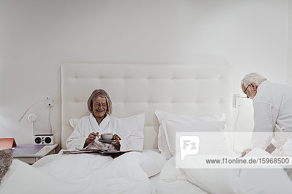 Glückliche ältere Frau trinkt Kaffee  während der Mann im Hotelzimmer am Bett steht