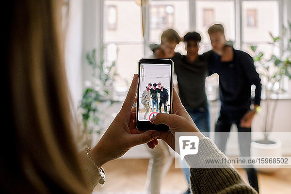 Teenager-Mädchen mit Smartphone filmt männliche Freunde  die mit dem Arm im Wohnzimmer herumstehen