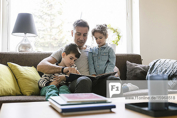 Father reading book to son and daughter while sitting on sofa at home