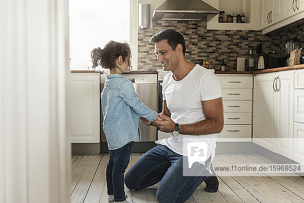 Smiling father holding hands with daughter while kneeling on kitchen floor