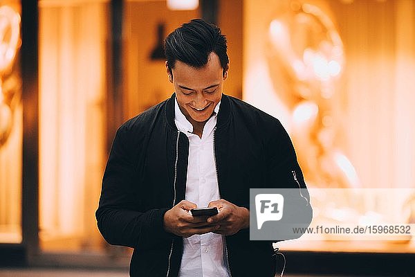 Lächelnder junger Mann benutzt Mobiltelefon  während er in beleuchteter Stadt steht