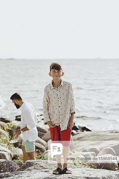 Portrait of smiling boy on rock against sea while father standing in background