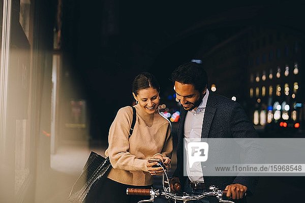 Smiling woman showing smart phone to male friend with bicycle while standing in city at night