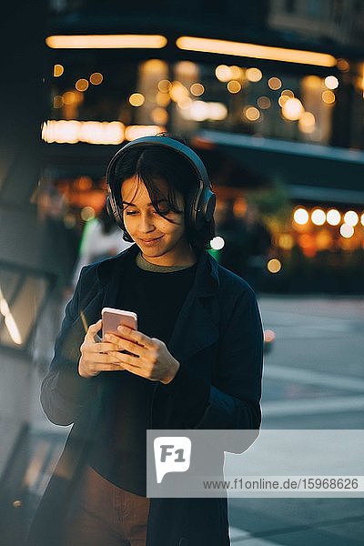 Junge Frau benutzt Mobiltelefon  während sie nachts in der Stadt Kopfhörer trägt