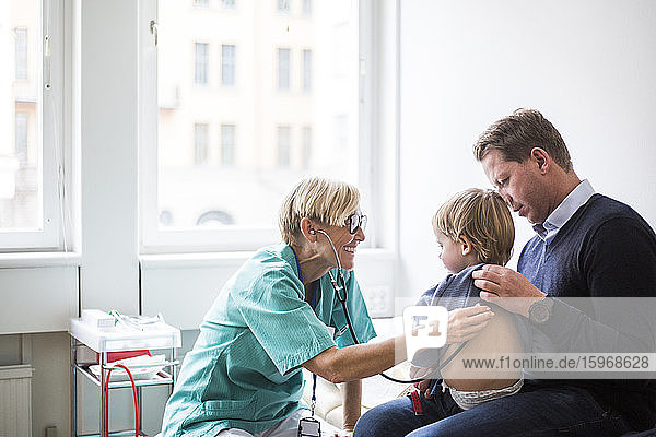 Lächelnde Ärztin untersucht den Rücken des Jungen mit dem Stethoskop  während sie im Krankenhaus beim Vater sitzt