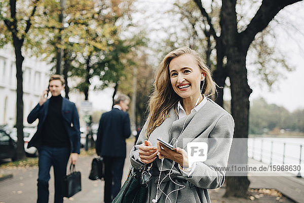 Lächelnde Geschäftsfrau mit In-Ohr-Kopfhörern und Smartphone in der Stadt stehend