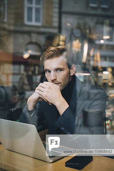 Portrait of businessman with laptop sitting in cafe seen through glass window