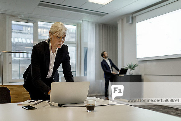 Geschäftsfrau benutzt Laptop  während ein Kollege während einer globalen Konferenzsitzung im Büro an einem Flachbildschirm steht