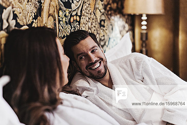 Happy mid adult man talking with girlfriend while relaxing on bed in hotel room