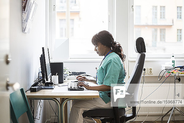 Seitenansicht einer reifen Ärztin  die über den Computer tippt  während sie in der Klinik sitzt