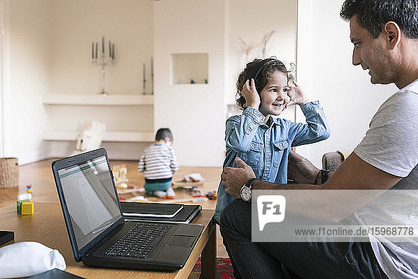 Lächelnde Tochter mit Kopfhörern steht neben dem Vater  während der Bruder zu Hause im Hintergrund spielt