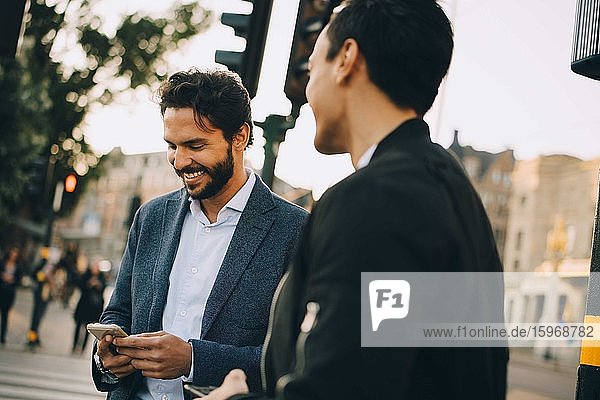 Smiling man using phone while standing by male friend in city