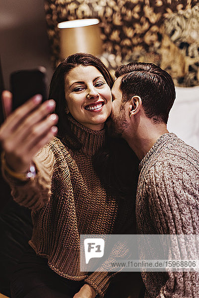 Man kissing while smiling girlfriend taking selfie in hotel room
