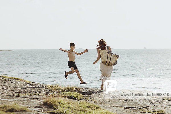 Rear view of woman with bag while son jumping against sea during sunny day