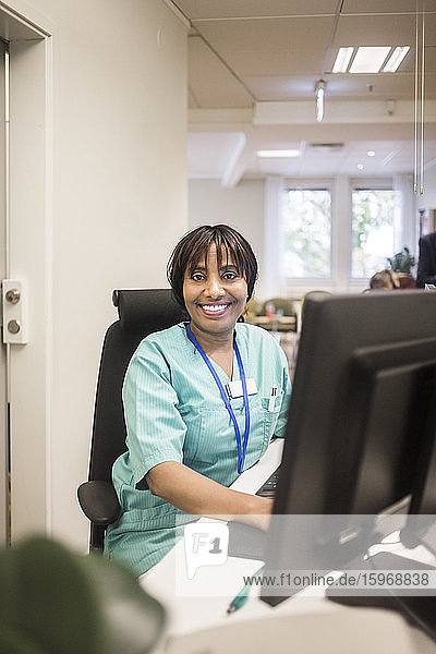 Porträt einer lächelnden Ärztin mit Ponyfrisur bei der Arbeit am Computer während sie in der Klinik sitzt