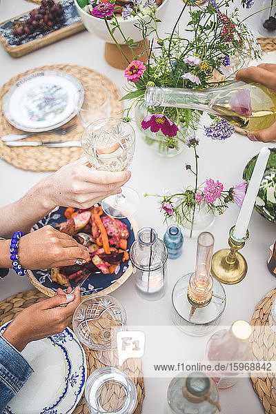 Schrägaufnahme einer geschorenen Hand  die Wein ausschenkt  während eine Frau während eines geselligen Beisammenseins auf einer Terrasse Essen serviert