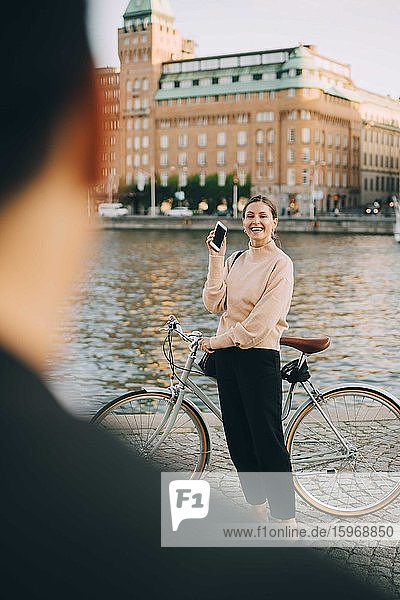 Lächelnde Frau mit Fahrrad schaut Freundin an  während sie am Fluss in der Stadt steht