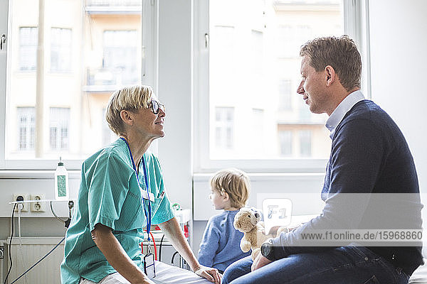 Seitenansicht einer reifen Ärztin  die mit einem Mann spricht  während ihr Sohn im Hintergrund in einer medizinischen Klinik steht