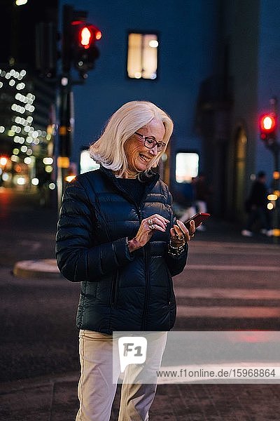 Lächelnde Frau schreibt eine SMS über ein Smartphone  während sie nachts in einer beleuchteten Stadt steht