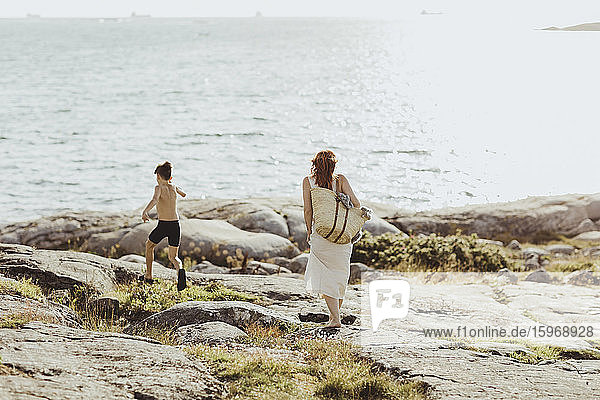Rückansicht einer Frau mit Tasche  während ihr Sohn bei Sonnenschein gegen das Meer läuft