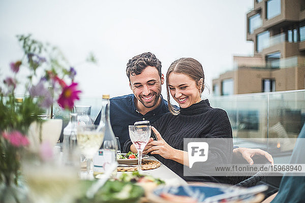 Glückliche männliche und weibliche Freunde verwenden ein Telefonfoto während einer Dachfeier