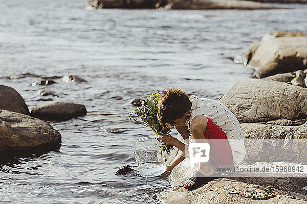 Junge kauert über Felsen  während er am Wochenende Wasser in Behälter füllt