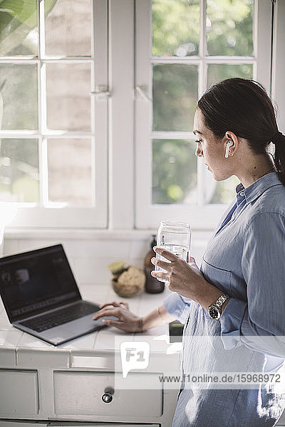 Seitenansicht eines weiblichen Mitarbeiters  der seinen Laptop am Tresen benutzt  während er im Heimbüro Wasser trinkt