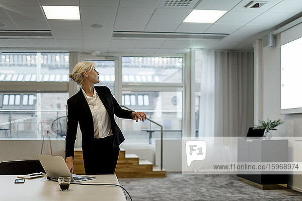 Selbstbewusste Geschäftsfrau benutzt Laptop  während sie im Sitzungssaal auf einen Flachbildschirm schaut