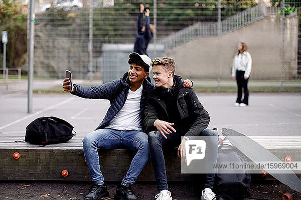 Teenager-Jungen in voller Länge  die mit dem Handy Selfie machen  während sie im Park sitzen