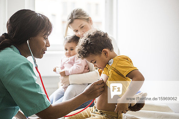 Seitenansicht einer Ärztin  die den Herzschlag des Jungen mit einem Stethoskop untersucht  während die Mutter sitzt und die Tochter in der Klinik ist