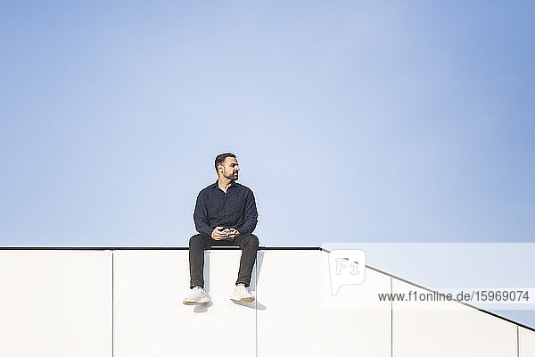 Kontemplierender Mann schaut weg  während er auf einem gebauten Gebäude vor blauem Himmel sitzt