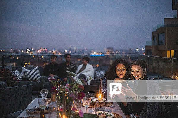 Lächelnde Frau zeigt einer Frau ein Smartphone  während Freunde im Hintergrund während eines geselligen Beisammenseins im Gebäude terr sitzen