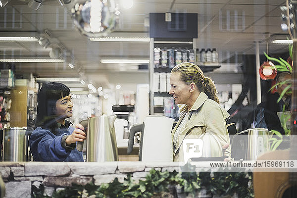 Verkäuferin mit Wasserkocher betrachtet Kundin im Geschäft durch Glasfenster gesehen