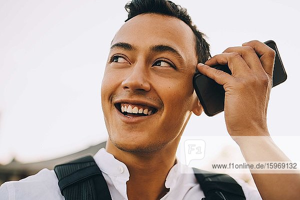 Niedrigwinkelansicht eines lächelnden jungen Mannes  der durch ein Smartphone gegen den Himmel lauscht