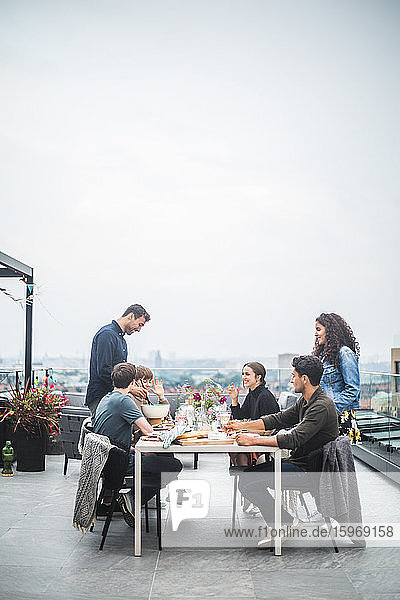 Smiling male with female serving food and drinks to friends during social gathering on rooftop