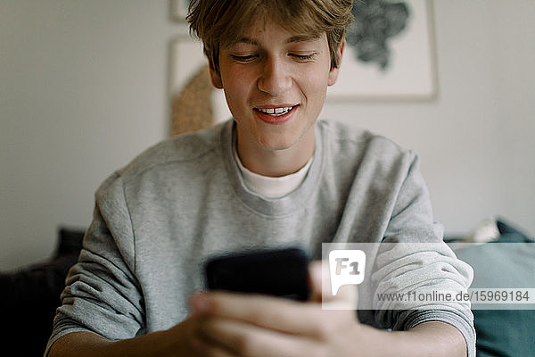 Smiling teenage boy using smart phone while sitting at home