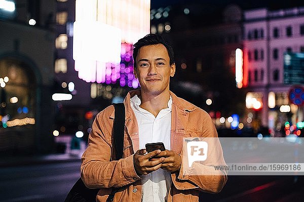 Portrait of smiling man using mobile phone while standing in illuminated city at night