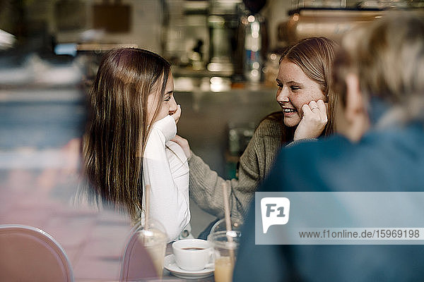 Smiling teenage female friends talking while sitting at cafe seen through glass window