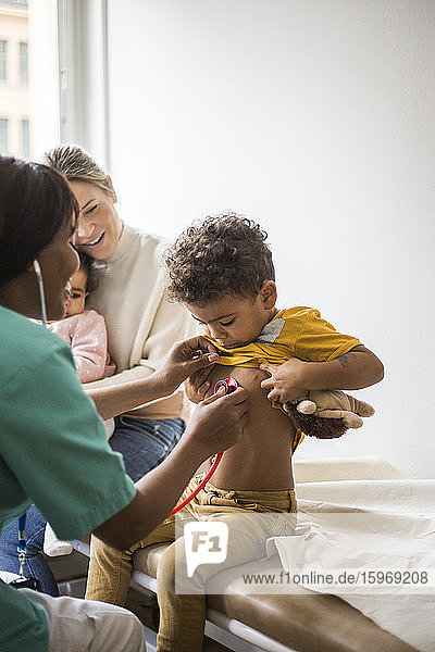 Weibliche Ärztin untersucht den Herzschlag des Jungen mit dem Stethoskop  während die Mutter sitzt und die Tochter in der Klinik