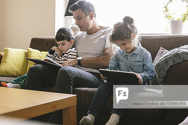 Vater liest dem Sohn ein Buch vor  während die Tochter ein digitales Tablett auf dem Sofa zu Hause benutzt
