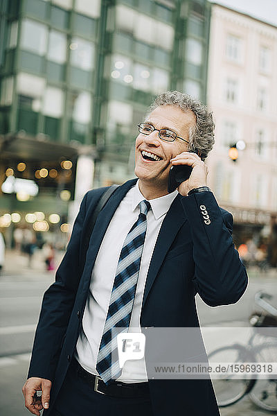 Lächelnder Geschäftsmann spricht durch ein Smartphone  während er in der Stadt steht