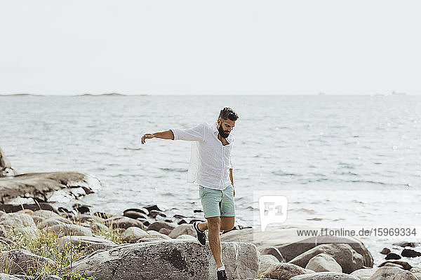 Man with arms outstretched walking over rocks against sea during sunny day