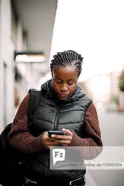 Teenage girl using smart phone while walking on footpath in city
