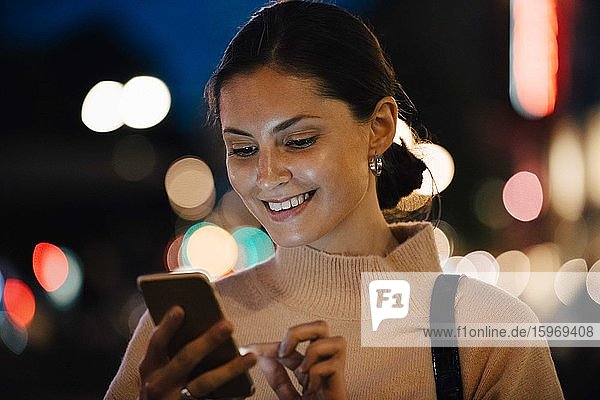 Lächelnde junge Frau schreibt nachts in der Stadt per Smartphone SMS