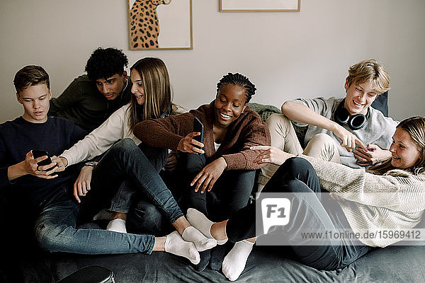 Lächelnde Teenager-Freunde nutzen soziale Medien auf Mobiltelefonen  während sie sich zu Hause auf dem Sofa entspannen